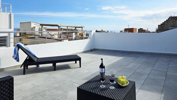 Una terraza en la azotea con una silla de descanso, una mesa con una botella y copas, y un bol de uvas.