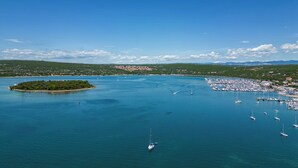 Vista aérea de um porto costeiro com barcos, uma pequena ilha e colinas ao longe.