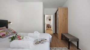 Quarto com cama, um banco, guarda-roupa de madeira e corredor que dá para outro cômodo.
