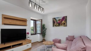 A modern living room with a flat-screen TV, a wooden shelf, a pink sofa, a small round table, and a painting of a street scene. - Seafront Apartment Nana 2 (Punat)