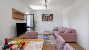 A modern living room with a flat-screen TV, a pink sofa, a wooden coffee table with a fruit bowl, and a painting on the wall. - Seafront Apartment Nana 2 (Punat)