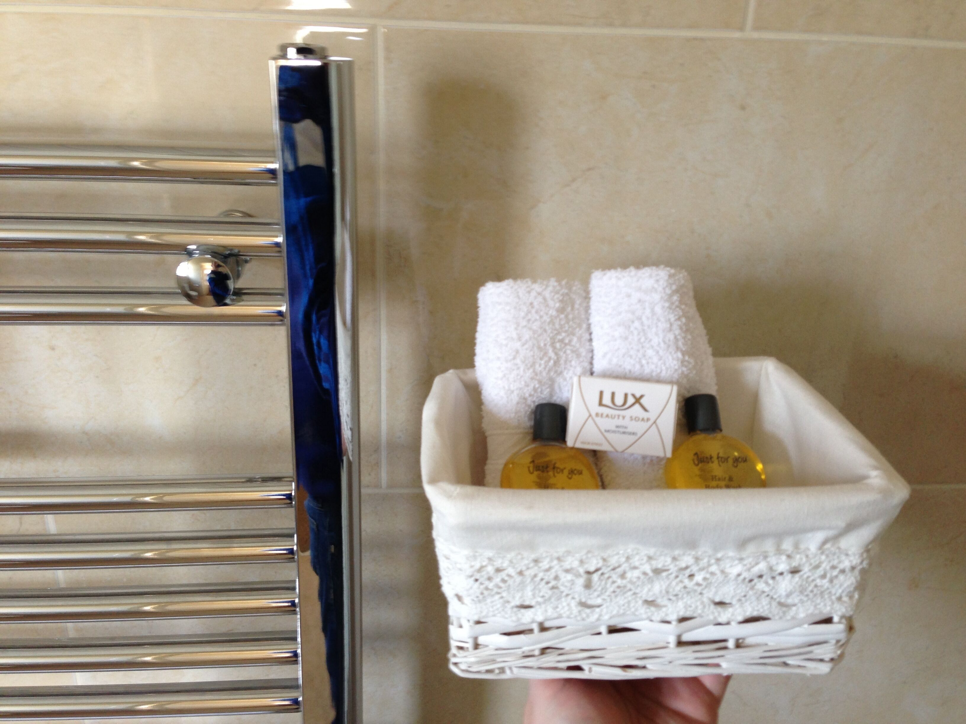 Une main tenant un panier en osier blanc contenant une boîte de savon de beauté Lux, deux flacons de produits de soins pour les cheveux et le corps, et deux serviettes blanches.
