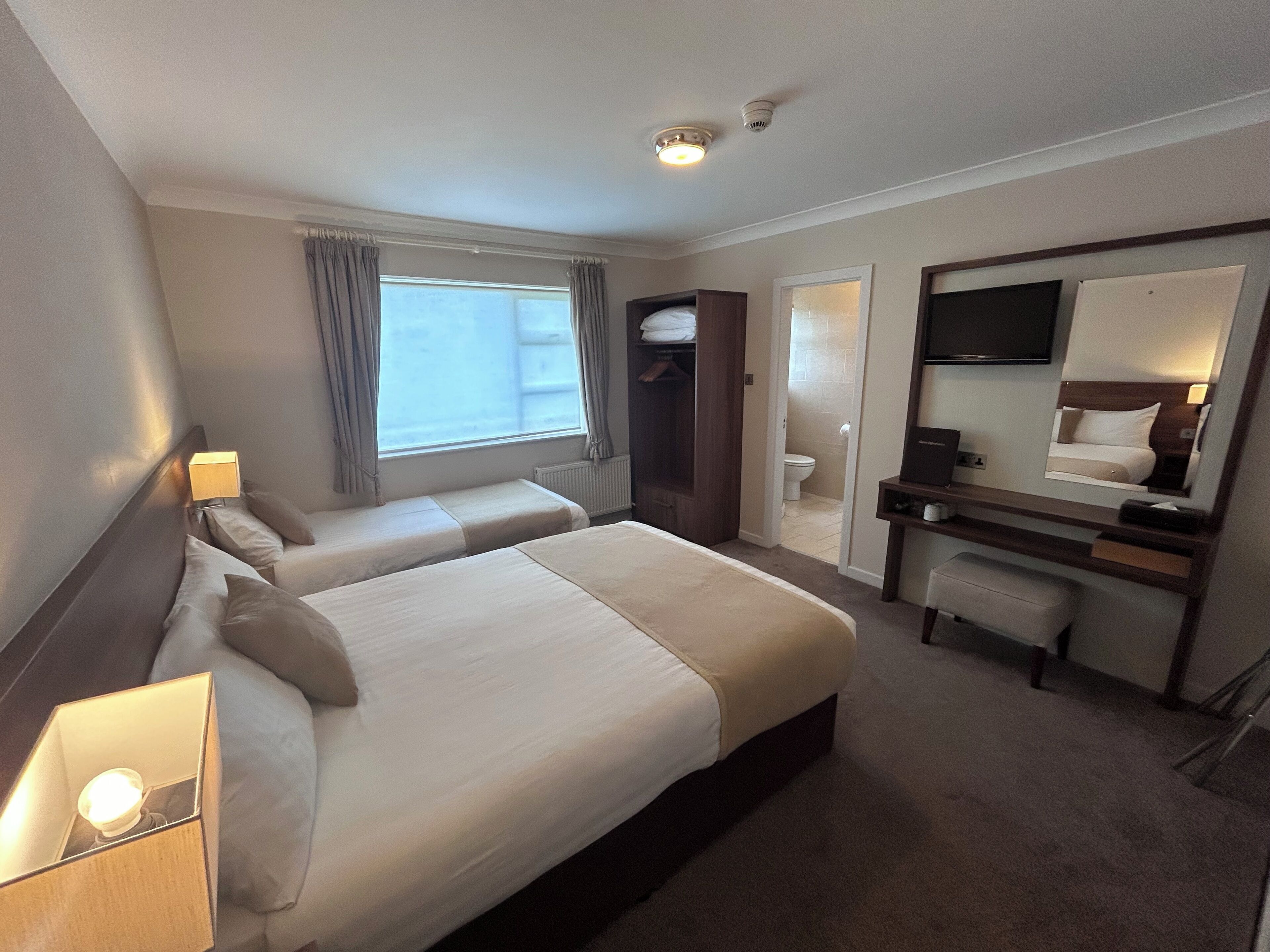 A hotel room with two beds, a desk, a mirror, and a bathroom visible through an open door.
