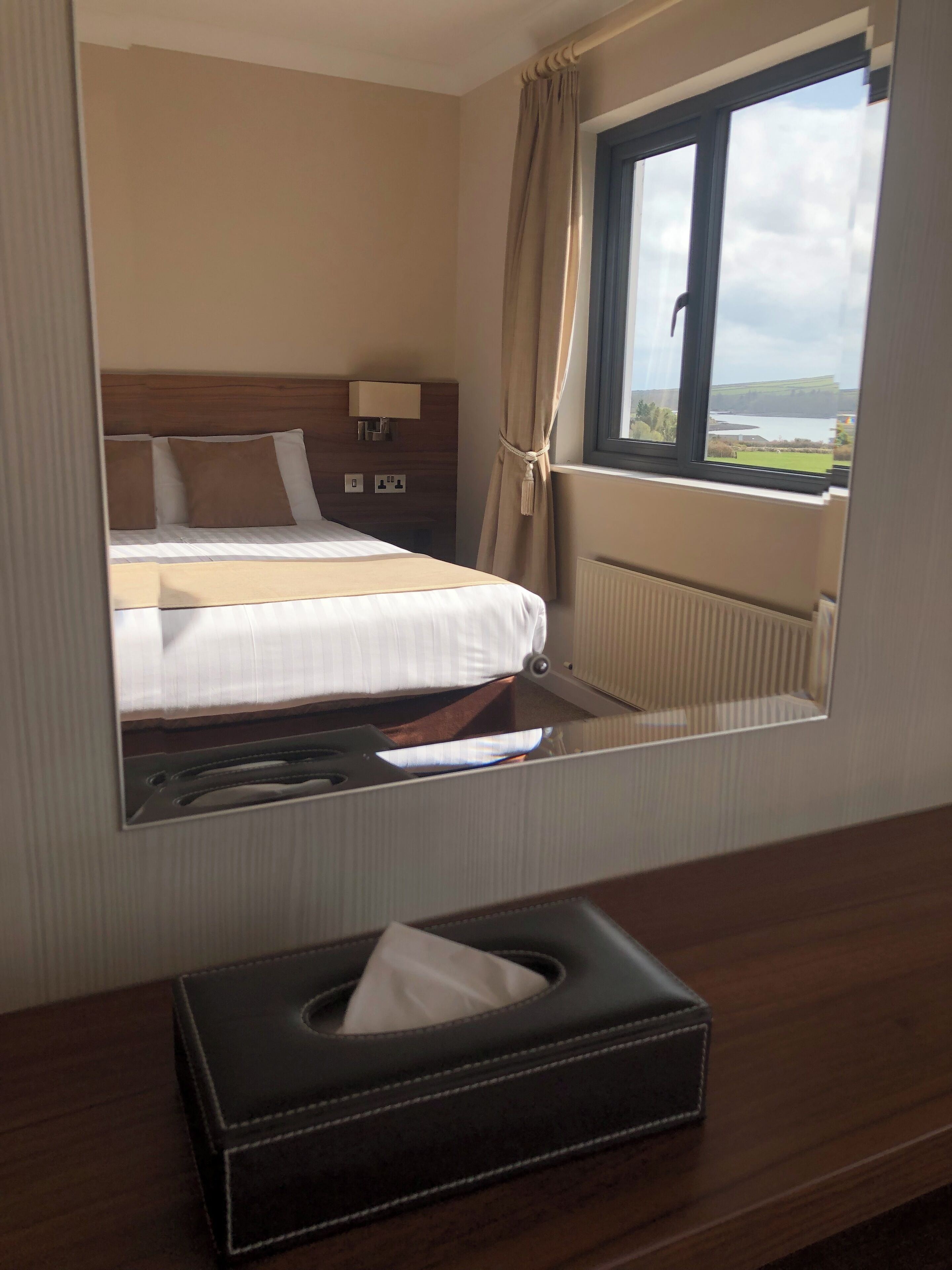 A hotel room with a bed, a bedside table with a lamp, a window with a view of a lake, and a mirror reflecting the room.