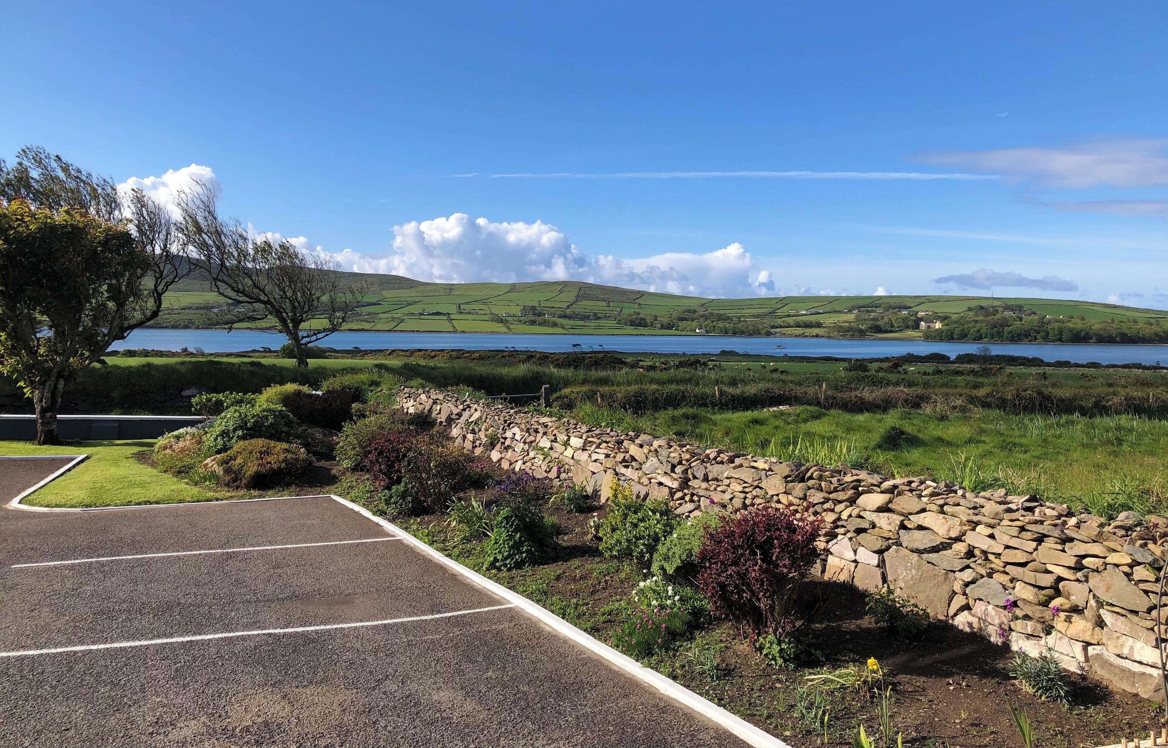 A parking area with a stone wall and greenery, leading to a scenic view of hills and a lake.