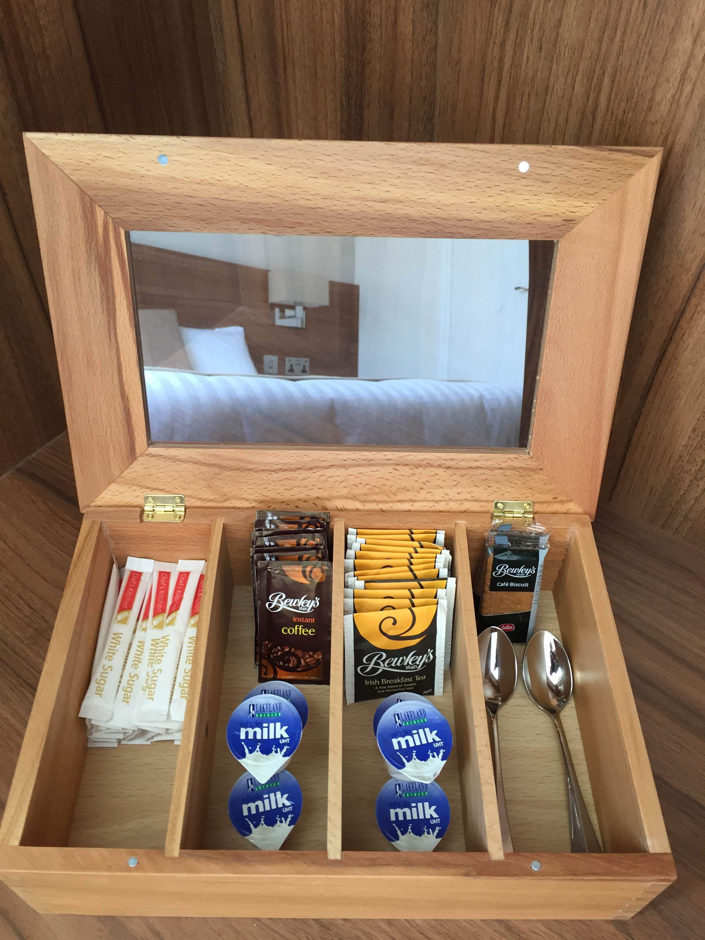 Un cofre de madera con compartimentos que contienen café instantáneo, té irlandés, sobres de azúcar blanca y cartones de leche.