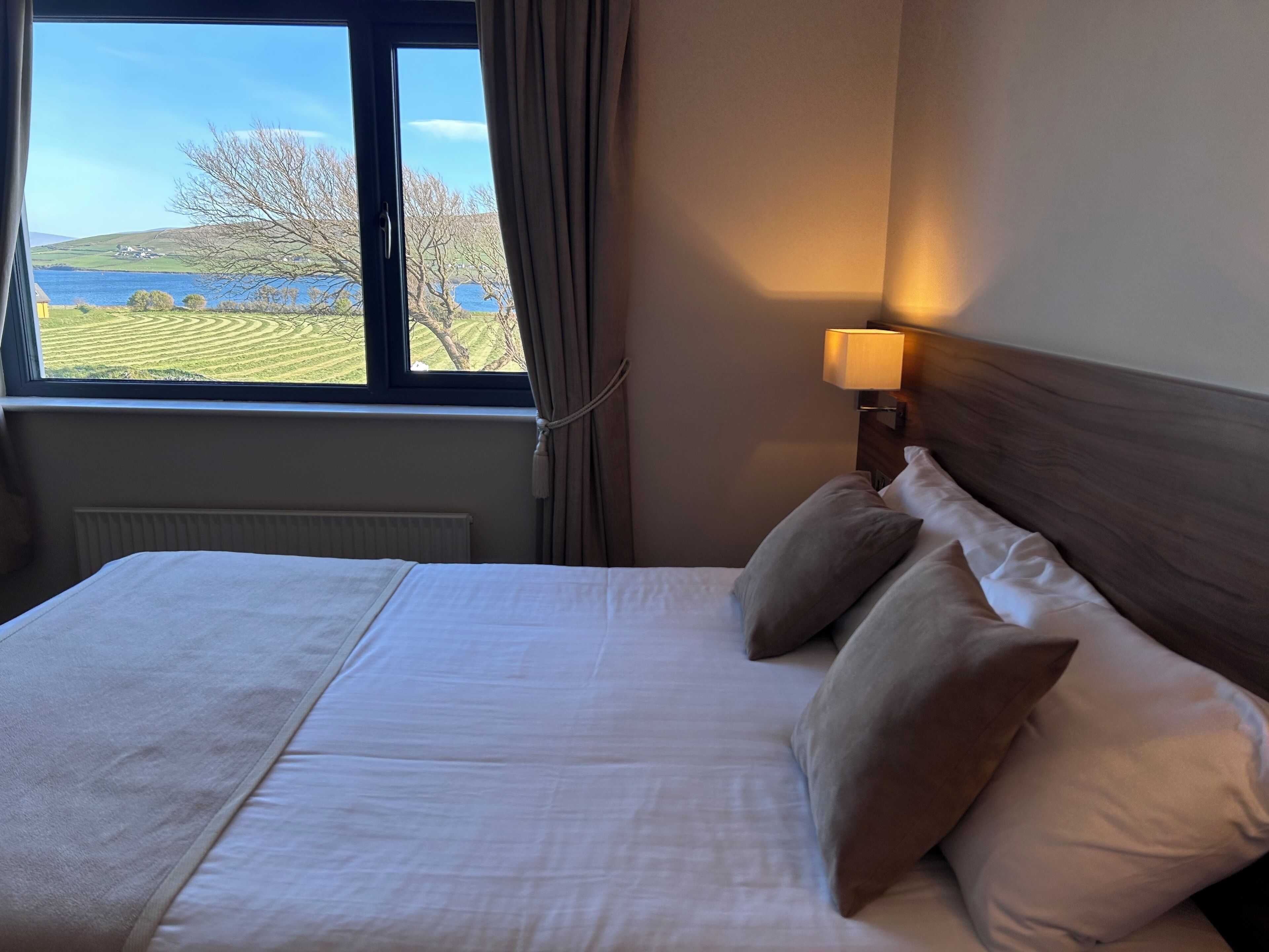 Ein Hotelzimmer mit einem großen Bett, einem Fenster mit Blick auf einen See und Hügel sowie einer Nachttischlampe.