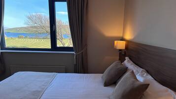 Ein Hotelzimmer mit einem großen Bett, einem Fenster mit Blick auf einen See und Hügel sowie einer Nachttischlampe.