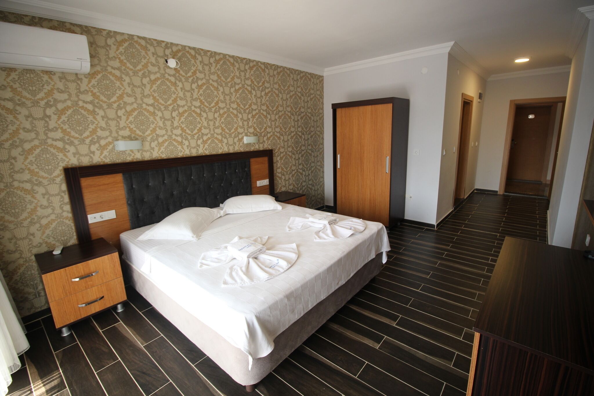 A hotel room with a bed, a wooden nightstand, a wardrobe, and a desk.