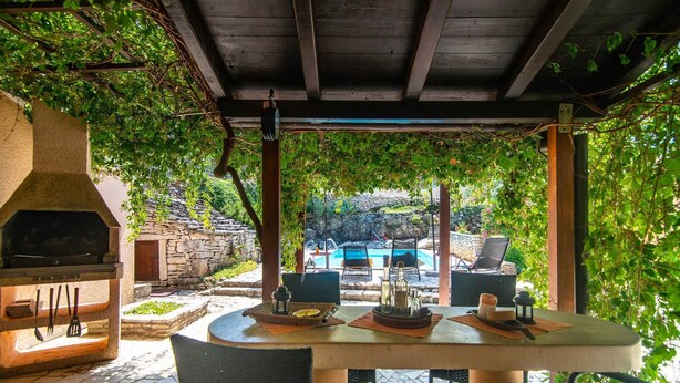 An outdoor patio with a stone barbecue, a table with chairs, and a view of a swimming pool. - Villa Giovanna (Rakalj)