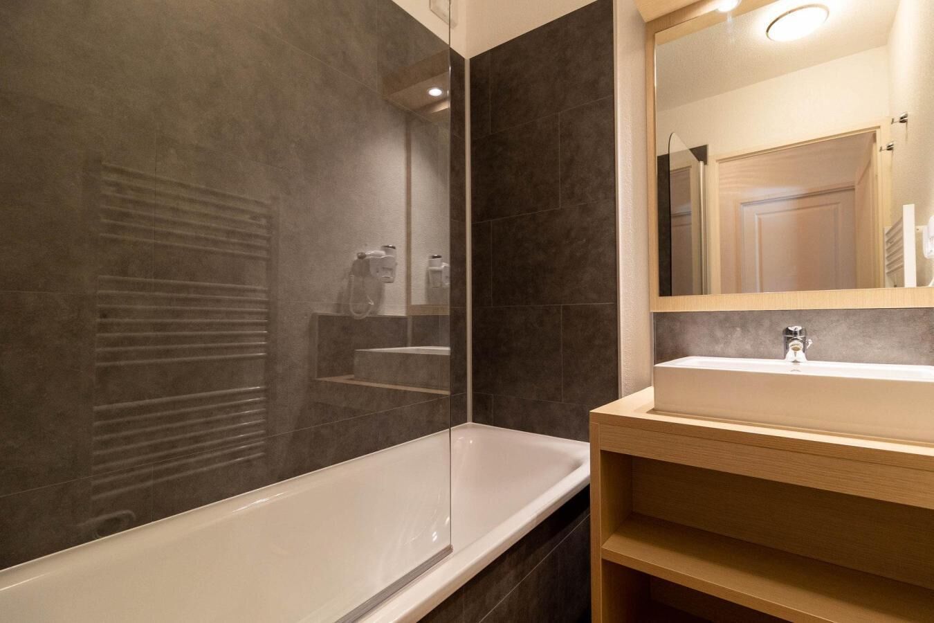 A modern bathroom with a bathtub, a vanity with a mirror, and dark tiled walls.