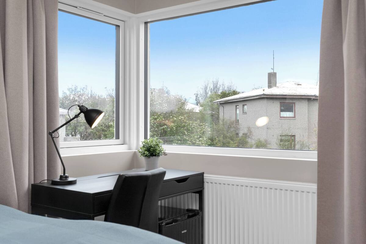 Un dormitorio con escritorio, lámpara, radiador y vista a un edificio a través de una ventana.