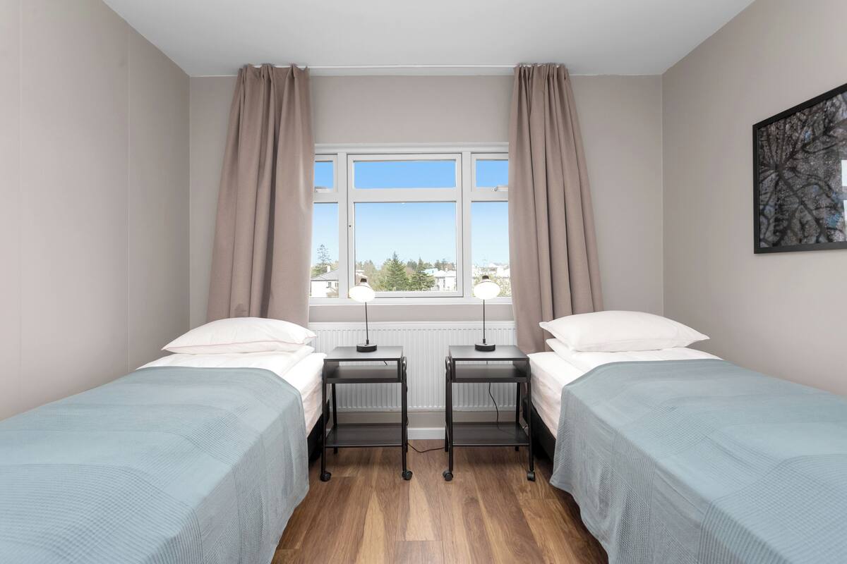 Habitación doble con dos camas individuales, una ventana con cortinas, un radiador y un cuadro enmarcado en la pared.