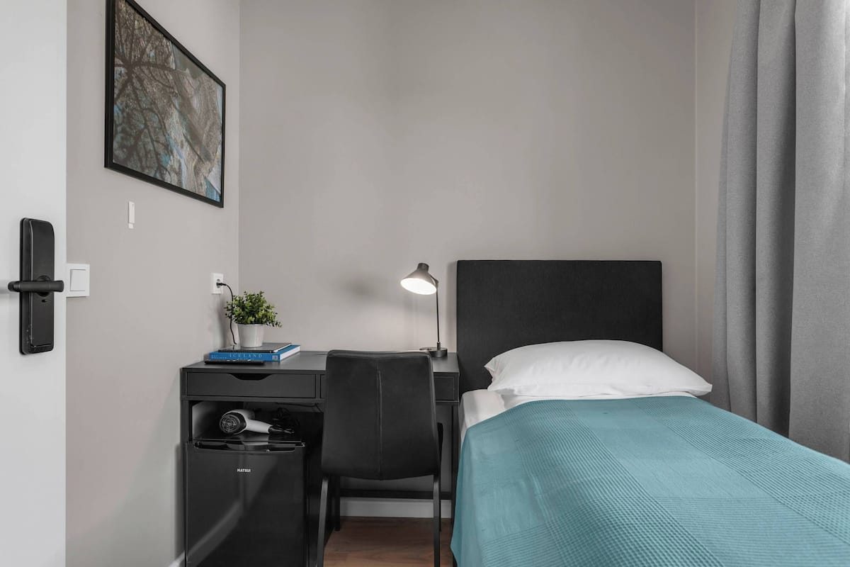 Habitación de hotel con cama, escritorio, silla y una pequeña mesa con lámpara y planta.
