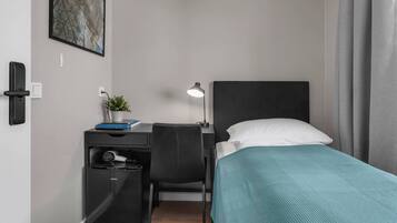 A hotel room with a bed, desk, chair, and a small desk with a lamp and a plant.