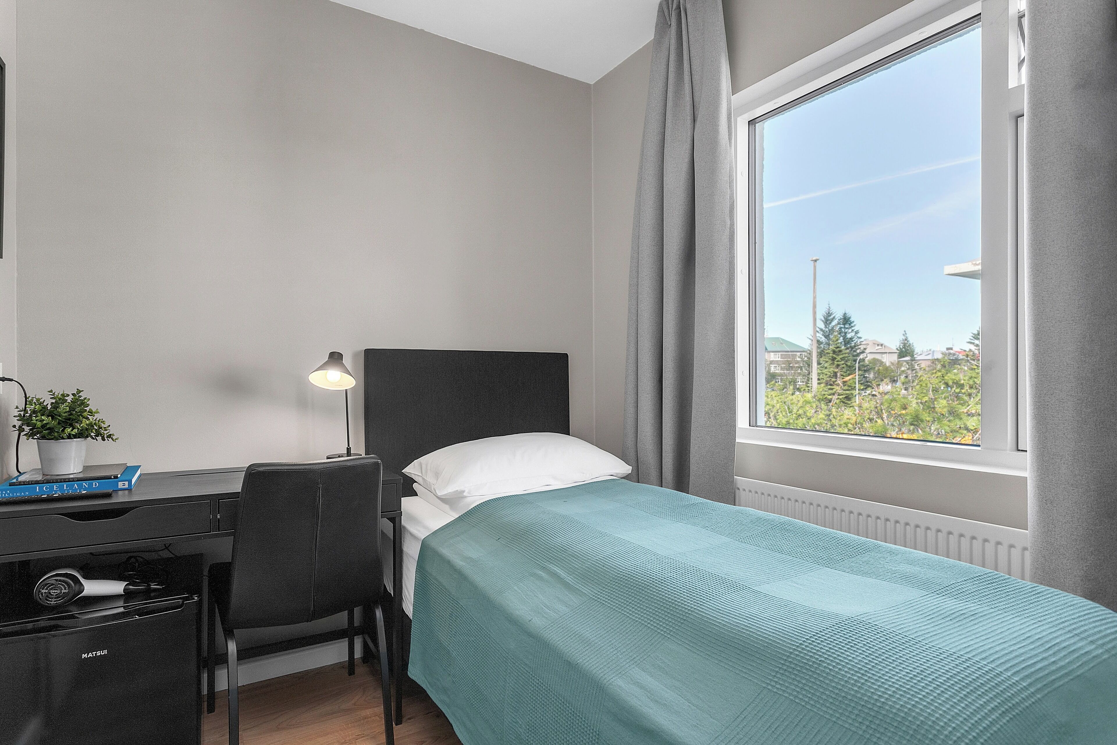 A hotel room with a bed, a desk, a chair, a lamp, a radiator, and a window with curtains.