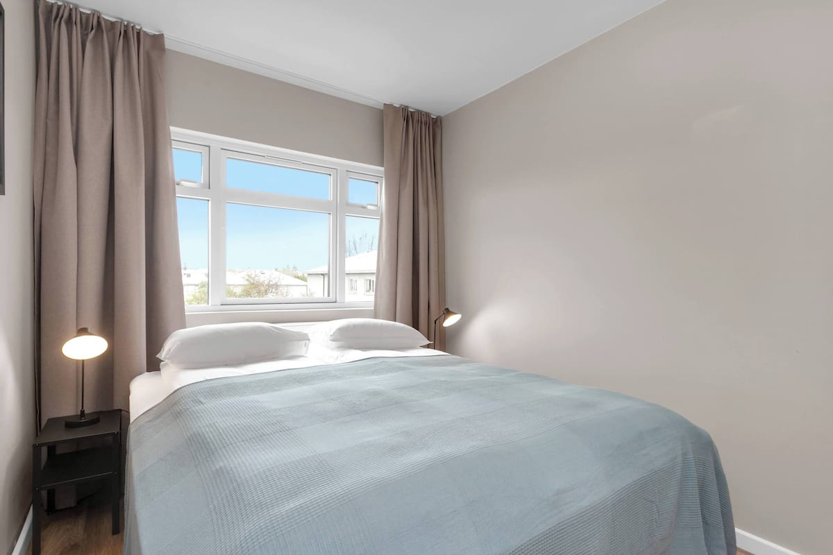 Un dormitorio con cama, dos lámparas de noche, ventana y cortinas.