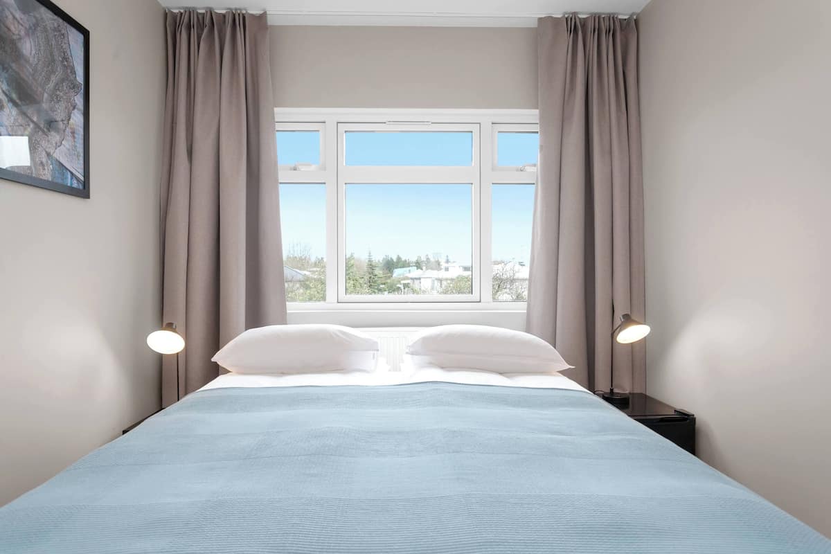 Un dormitorio con cama, dos lámparas de noche, una ventana con cortinas y un cuadro en la pared.