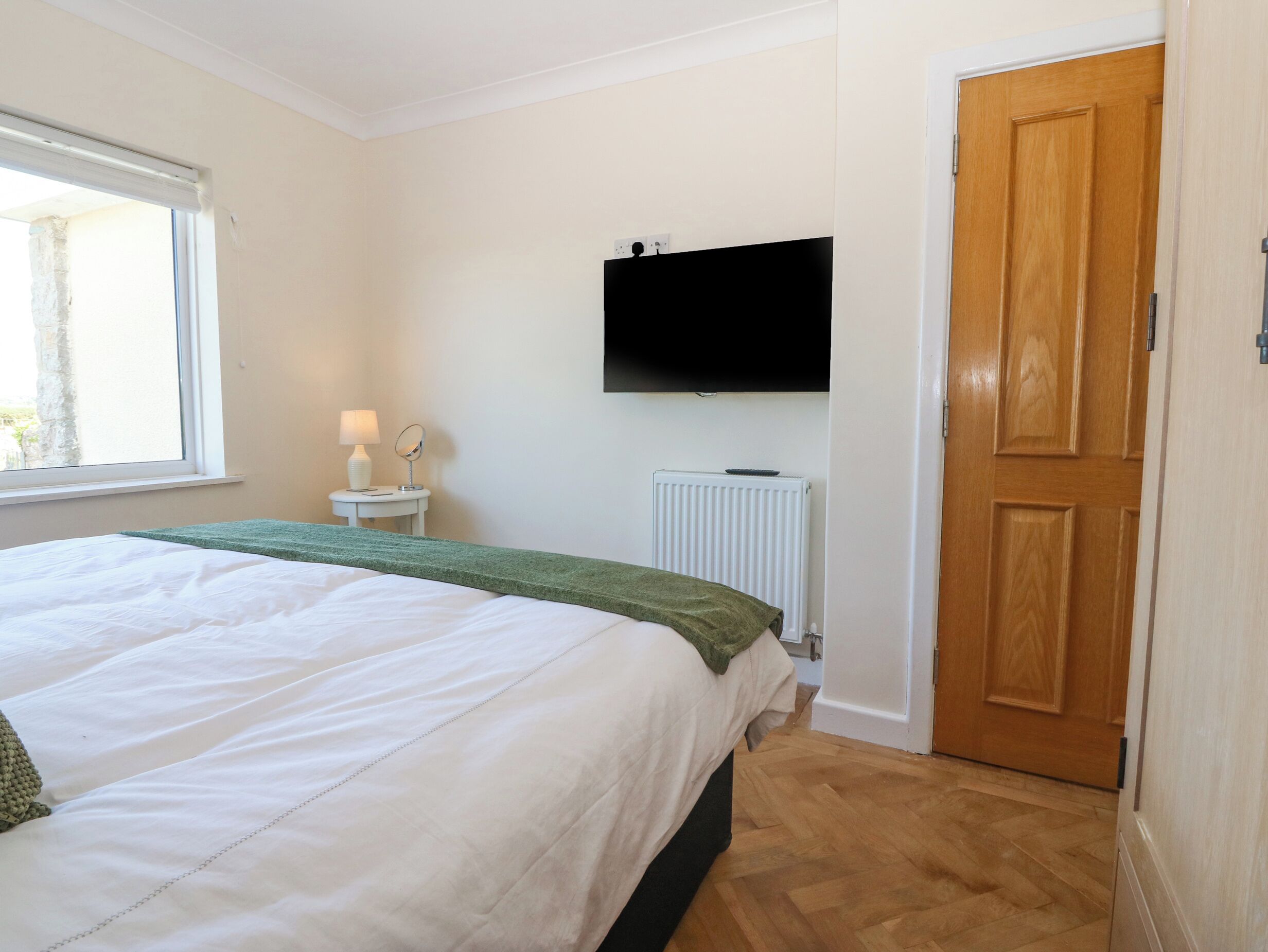 A bedroom with a bed, a bedside table with a lamp, a television mounted on the wall, a radiator, and a wooden door.