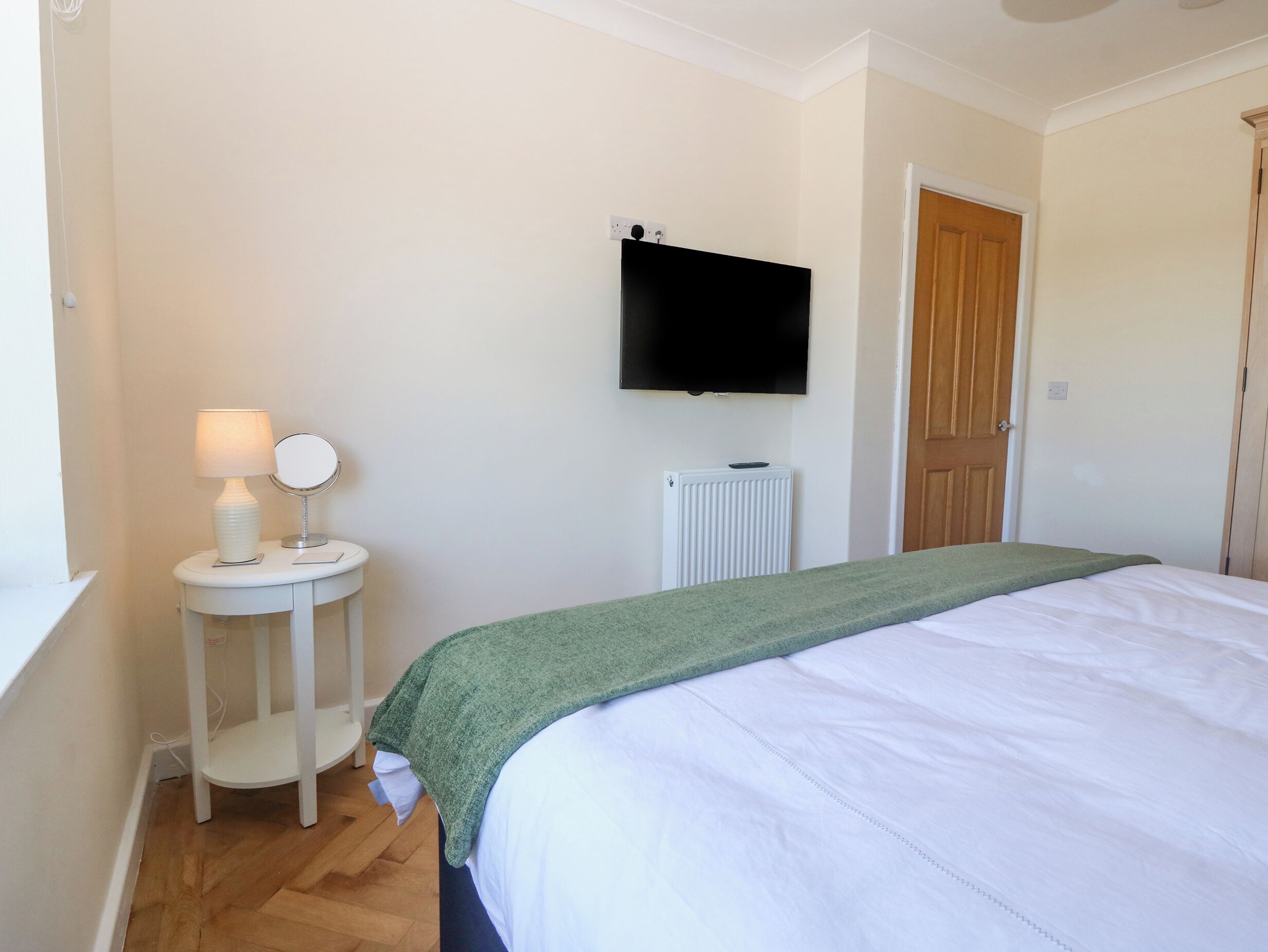 A bedroom with a bed, a bedside table with a lamp, a mirror, a wall-mounted TV, and a radiator.