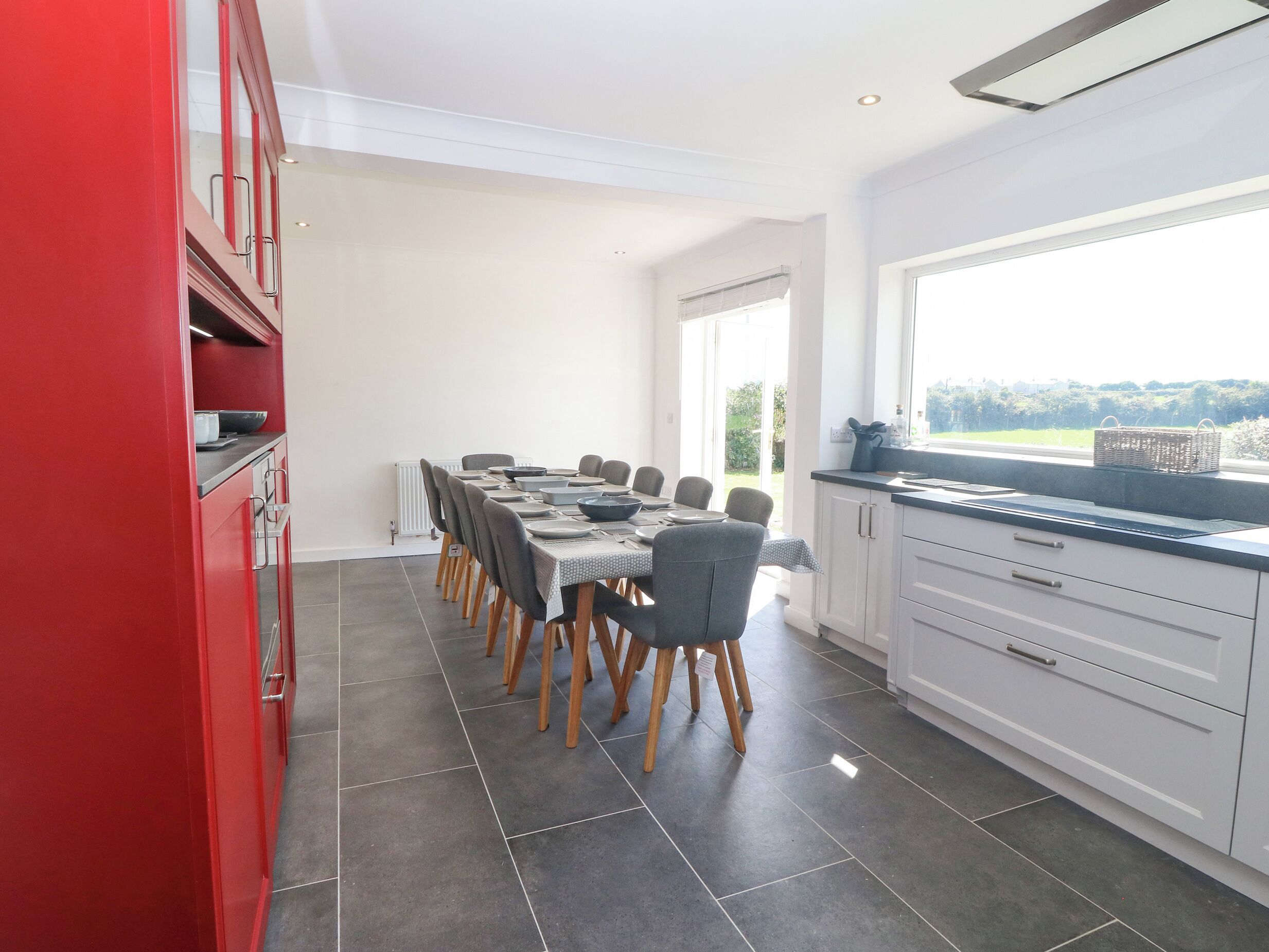 A modern kitchen with a dining area, featuring a long table, chairs, and a view of the outdoors through a large window.
