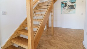Un escalier en bois avec des rampes en verre, dans une pièce dotée d’un parquet en bois disposé en chevrons et d’un tableau représentant une vache accroché au mur.