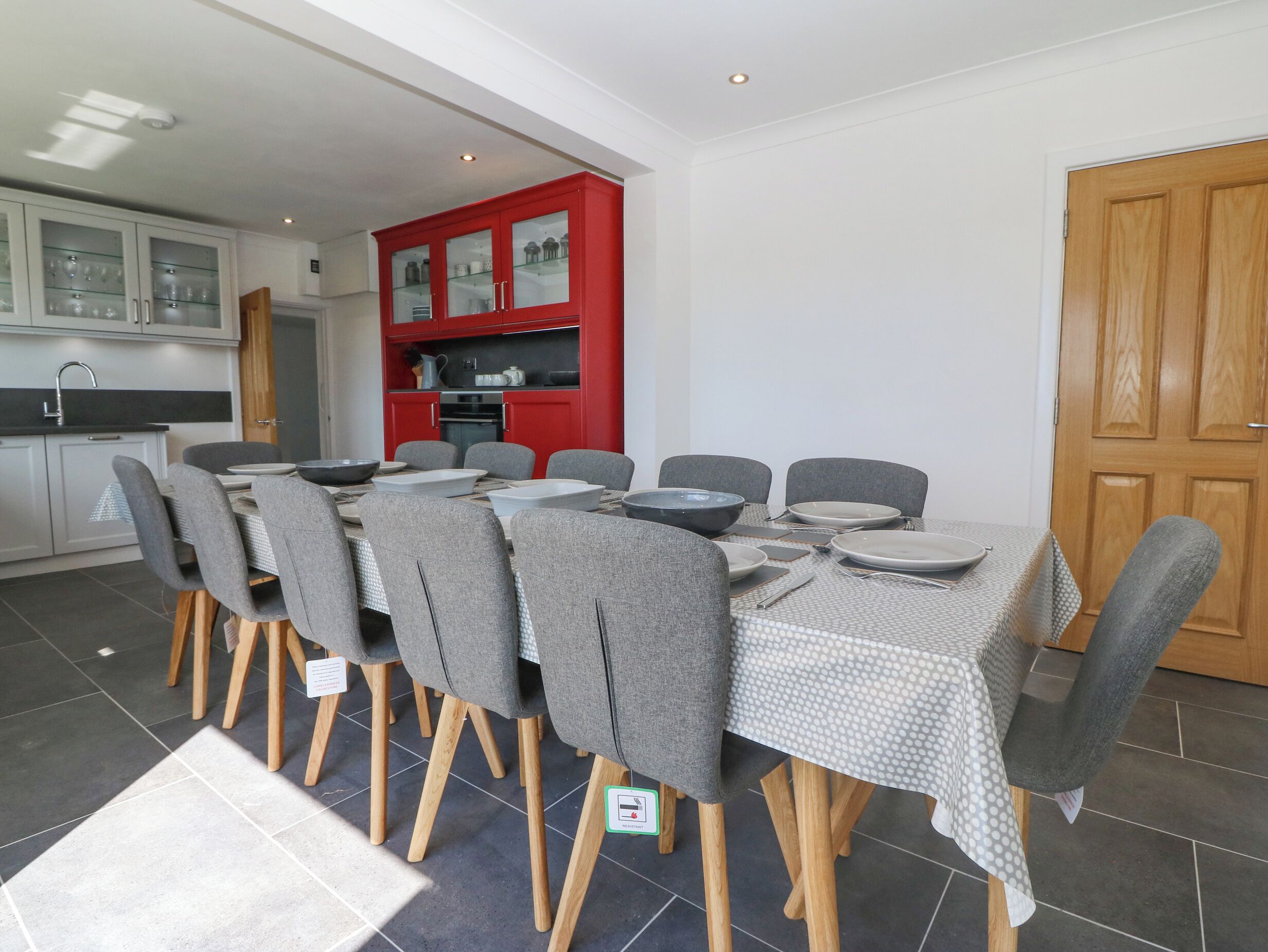 A dining area with a table set for six, surrounded by grey chairs with wooden legs. A red cabinet with glass doors is visible in the background.