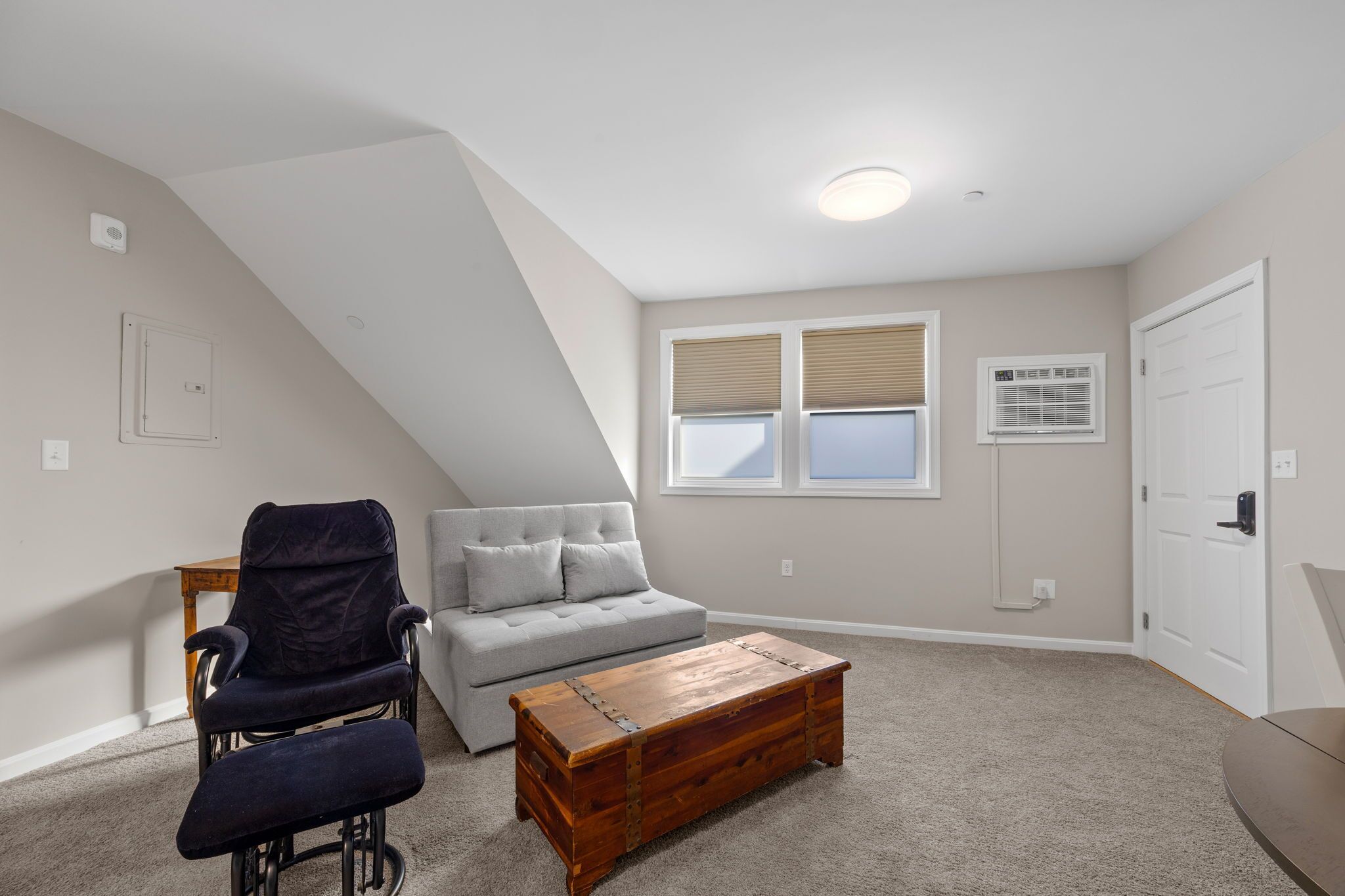 Une chambre à coucher aménagée dans un espace mansardé avec un plafond en pente, un canapé gris, un fauteuil foncé, une table basse en bois et une fenêtre avec des stores.