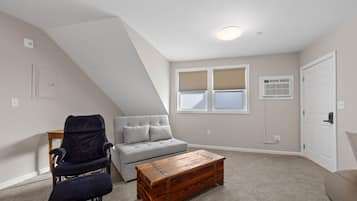 Une chambre à coucher aménagée dans un espace mansardé avec un plafond en pente, un canapé gris, un fauteuil foncé, une table basse en bois et une fenêtre avec des stores.