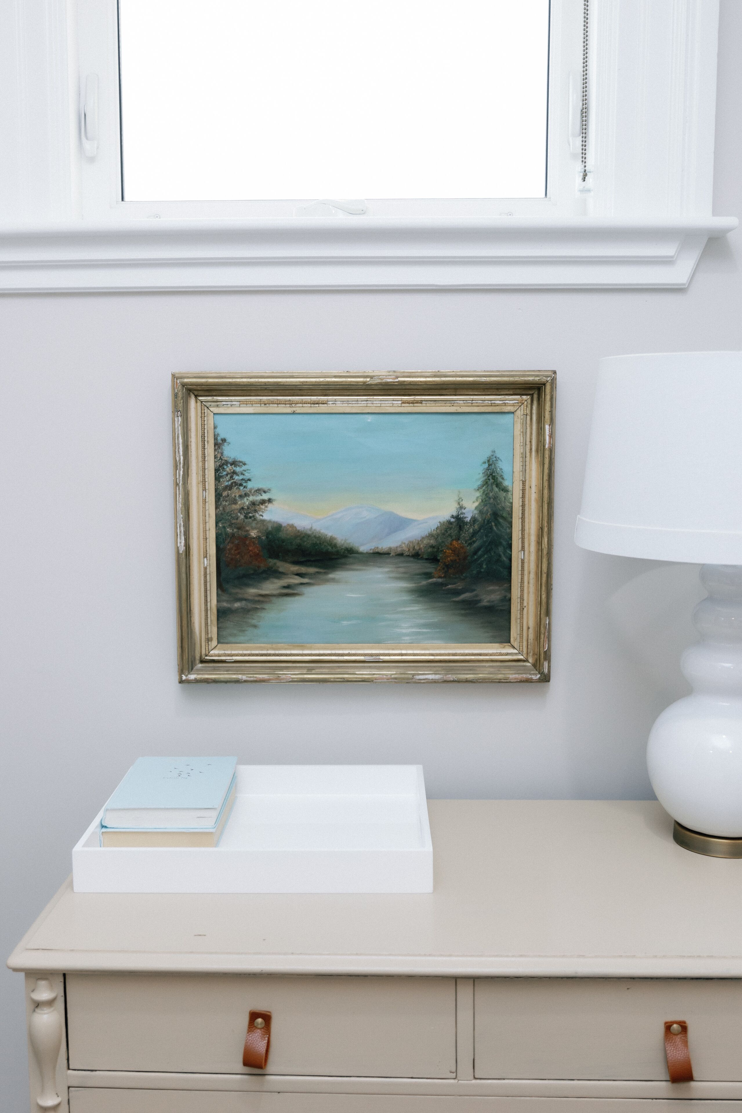 A room with a white cabinet above a desk, a framed painting on the wall, and a white lamp beside the desk.