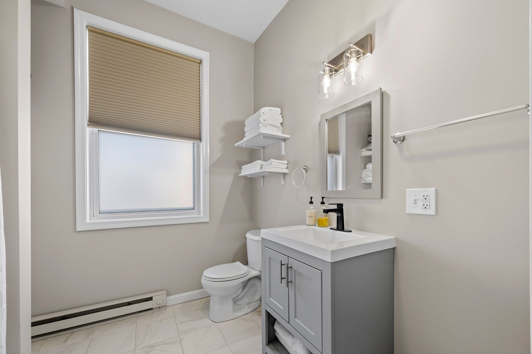 A bathroom with a toilet, sink, mirror, towel rack, and window with blinds.