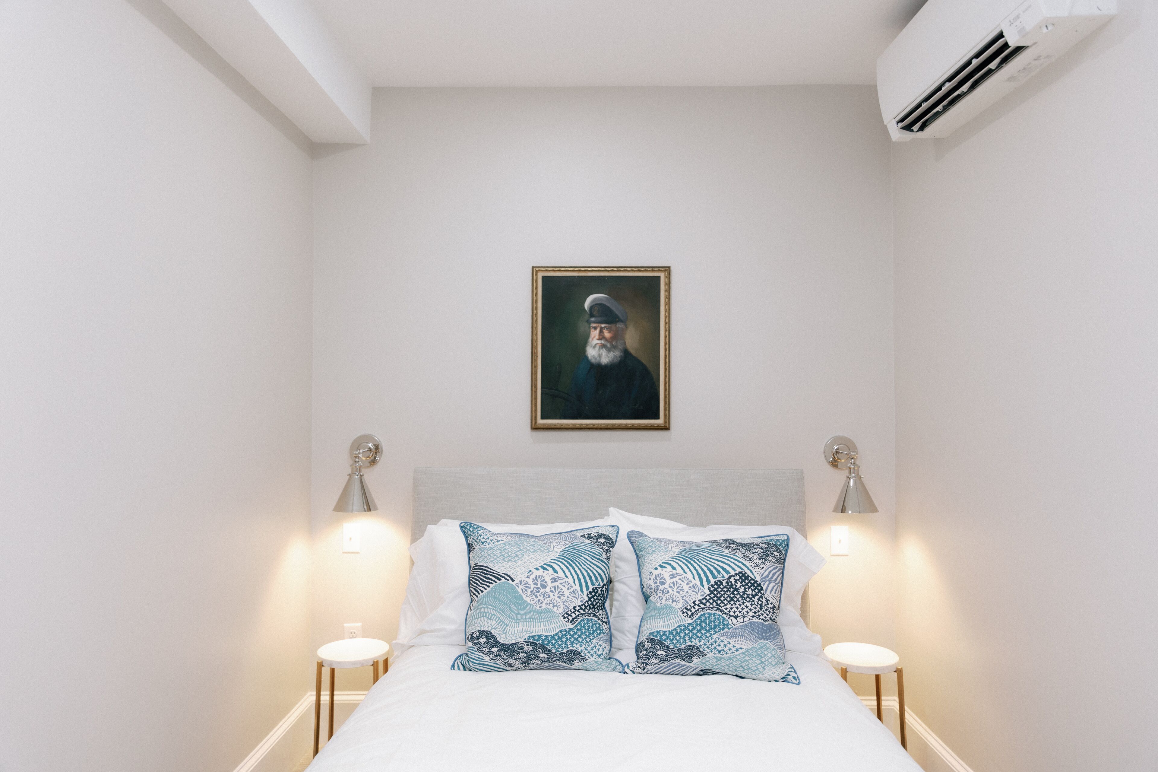 A bedroom with a bed, two bedside tables, a wall-mounted air conditioner, and a framed portrait above the bed.