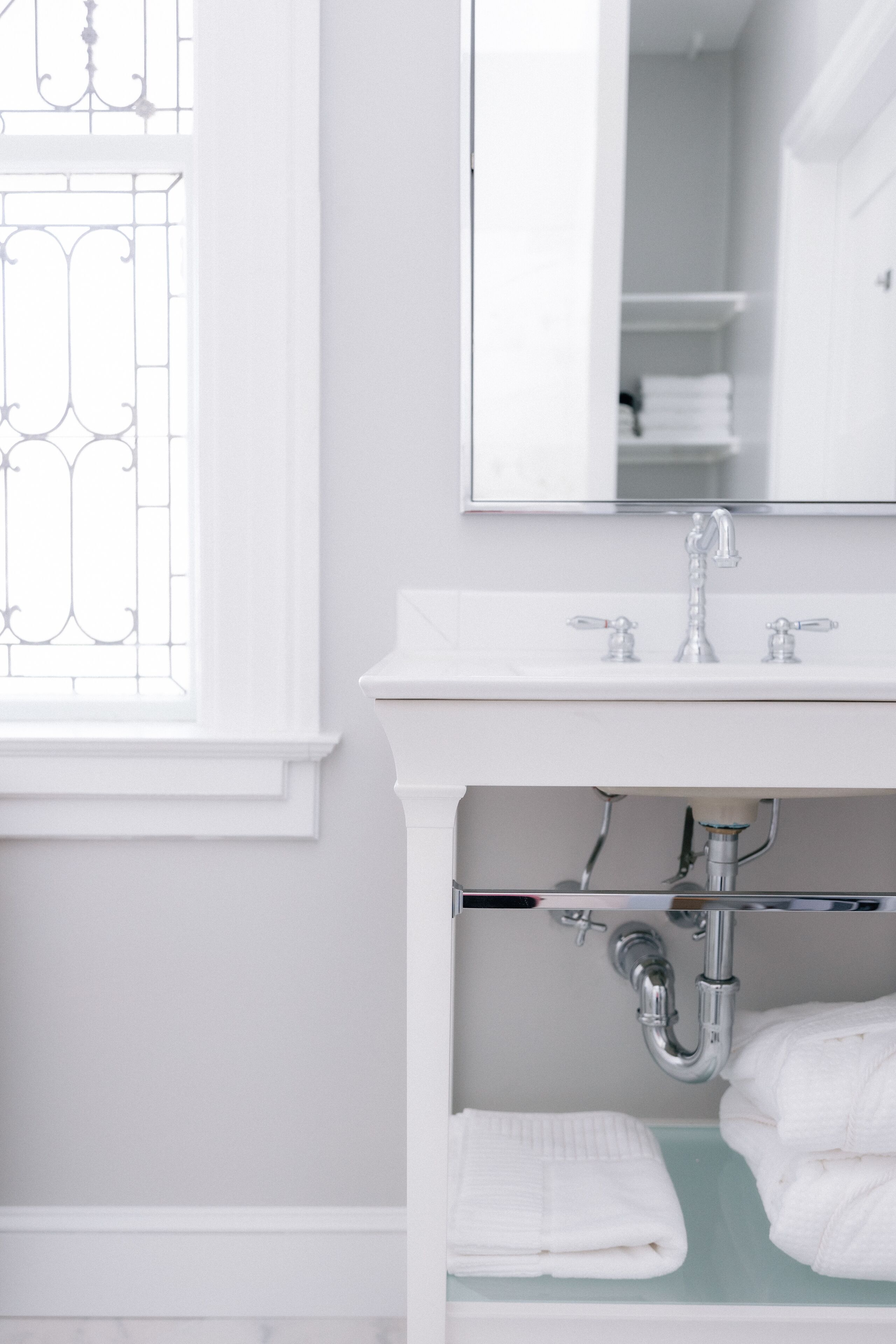 Een badkamer met een wit wastafelmeubel, een spiegel erboven en een raam met decoratief glaswerk.