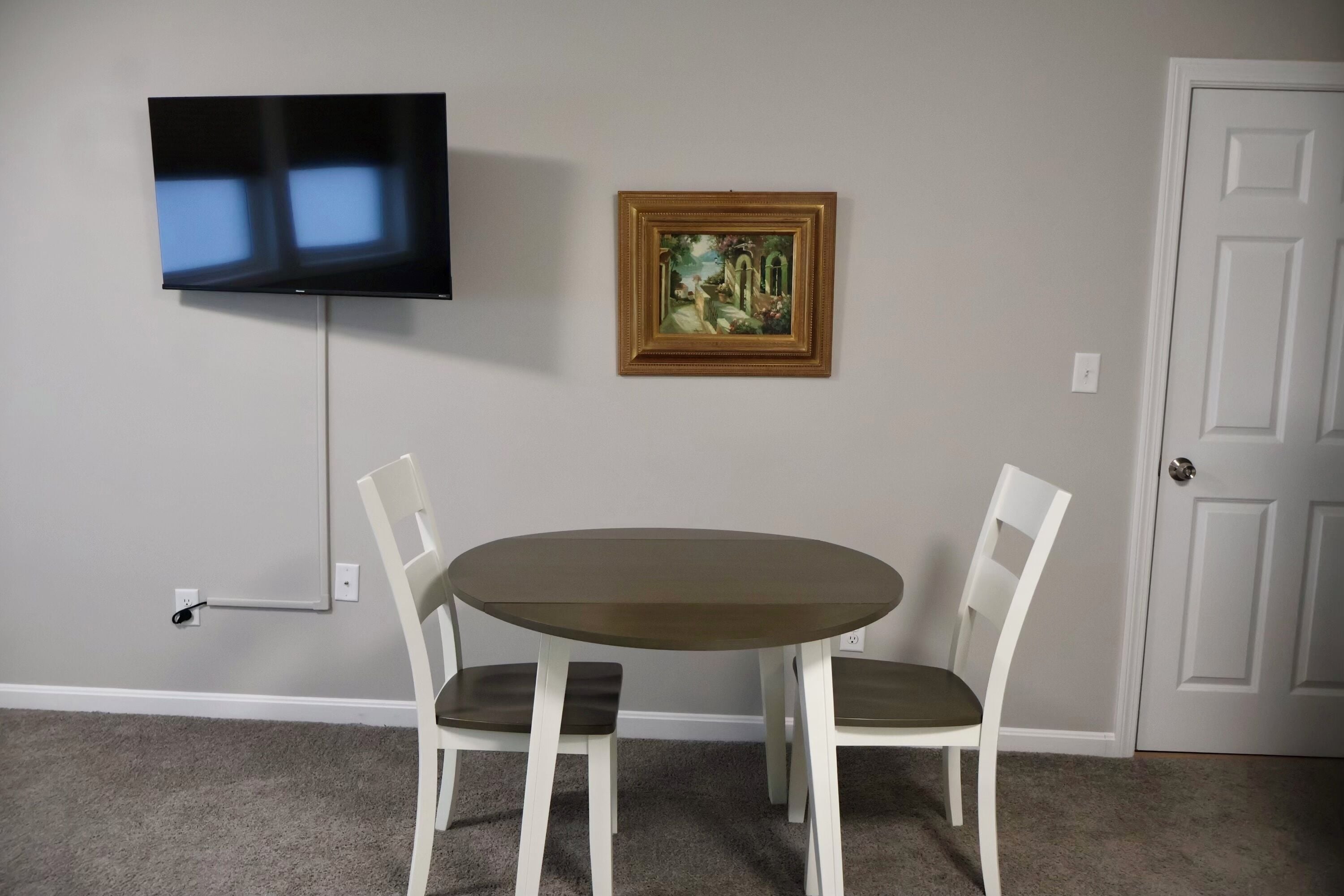 Une pièce avec un téléviseur à écran plat fixé au mur, un tableau encadré au-dessus d’une table ronde accompagnée de deux chaises, et une porte blanche sur la droite.