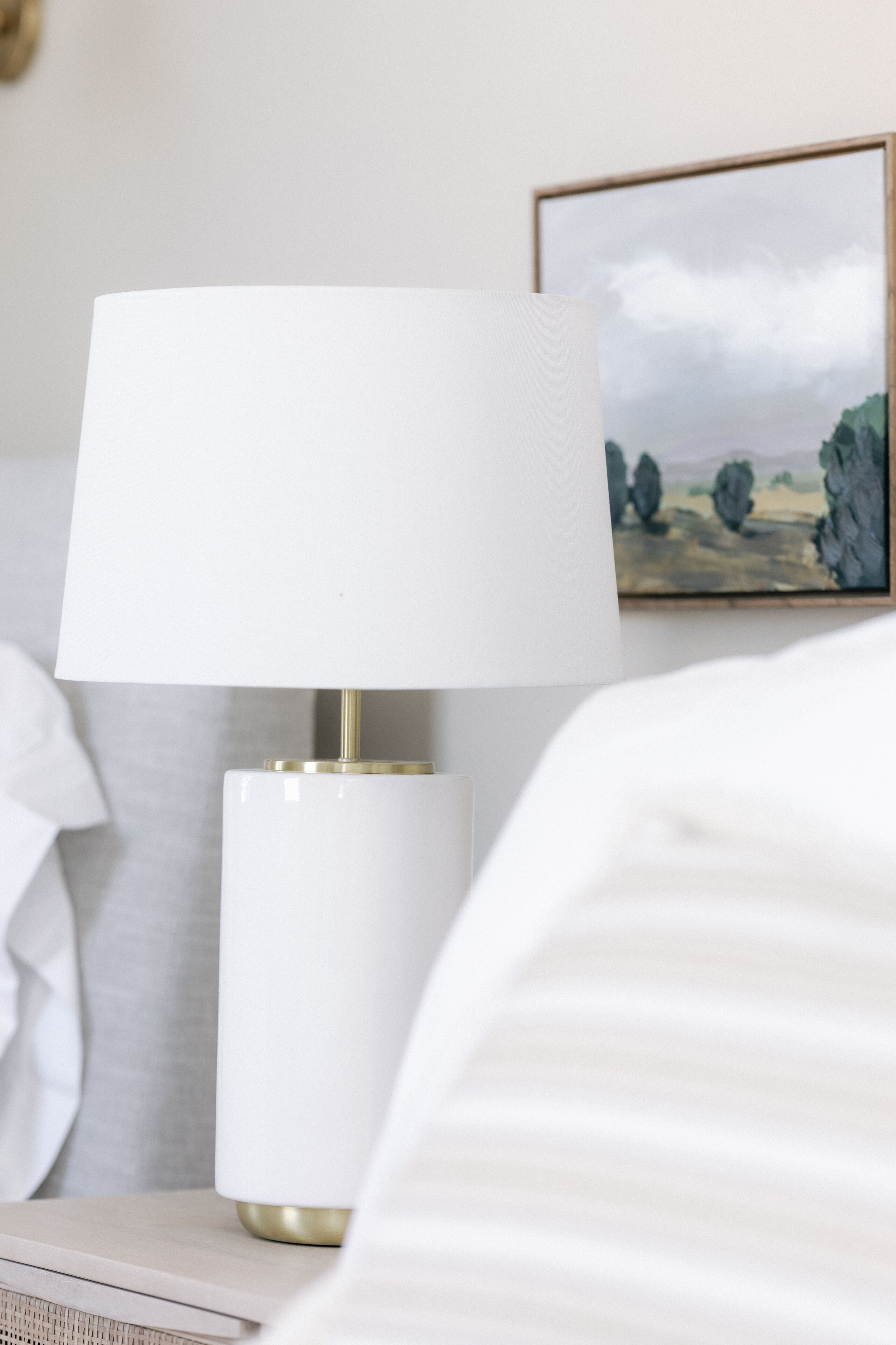 A bedside table with a white lamp and a framed landscape painting.