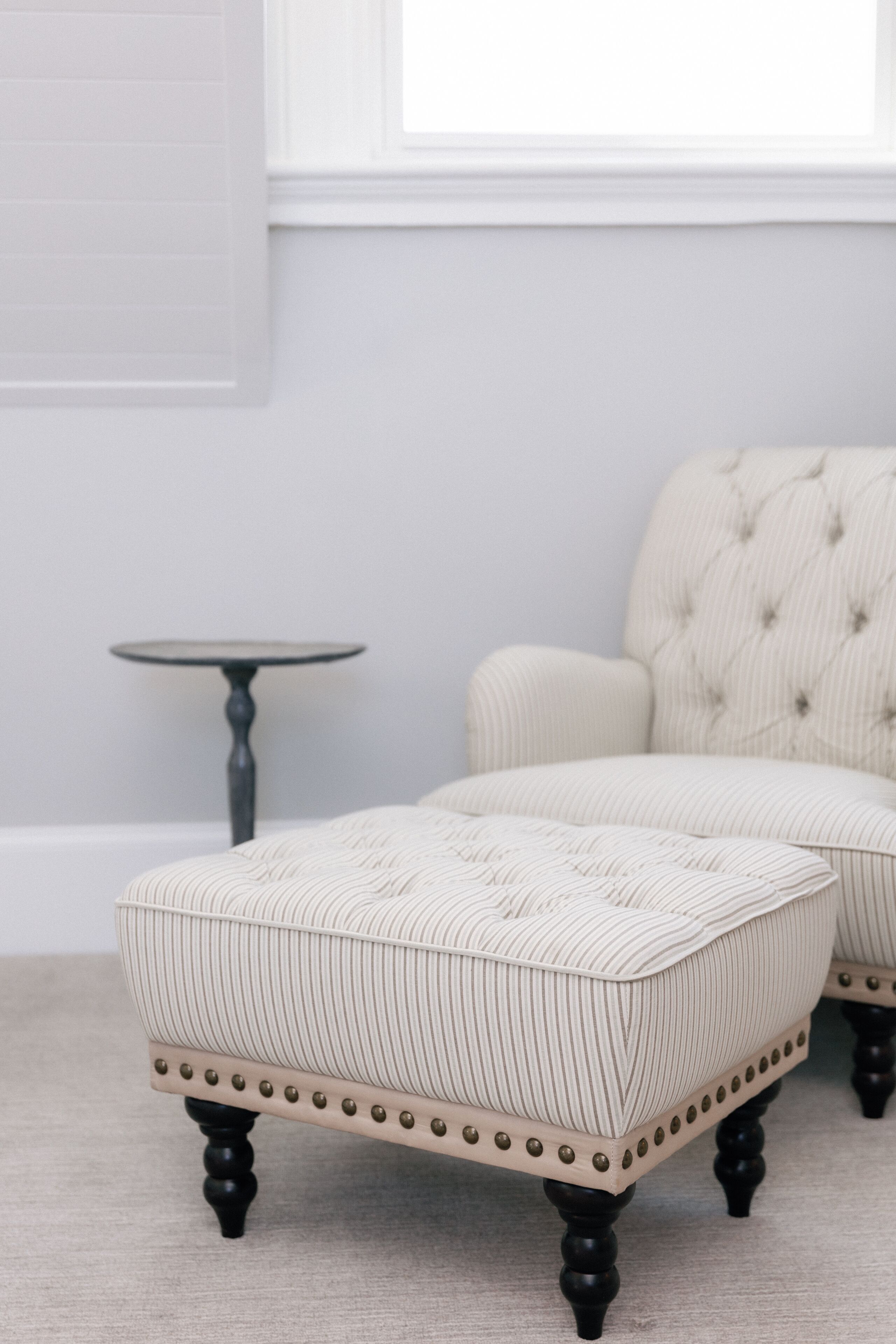 A room with a tufted armchair and ottoman, a small round table, and a window with blinds.