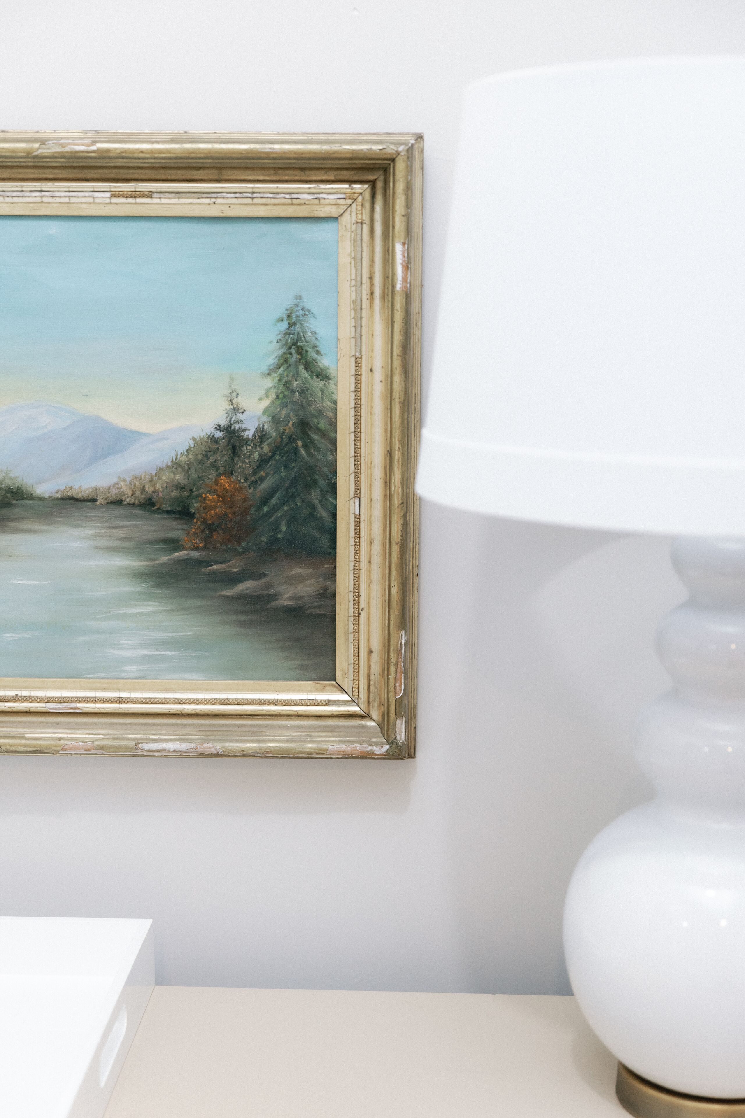 Un cuadro de paisaje enmarcado en la pared, una mesa blanca y una lámpara blanca.