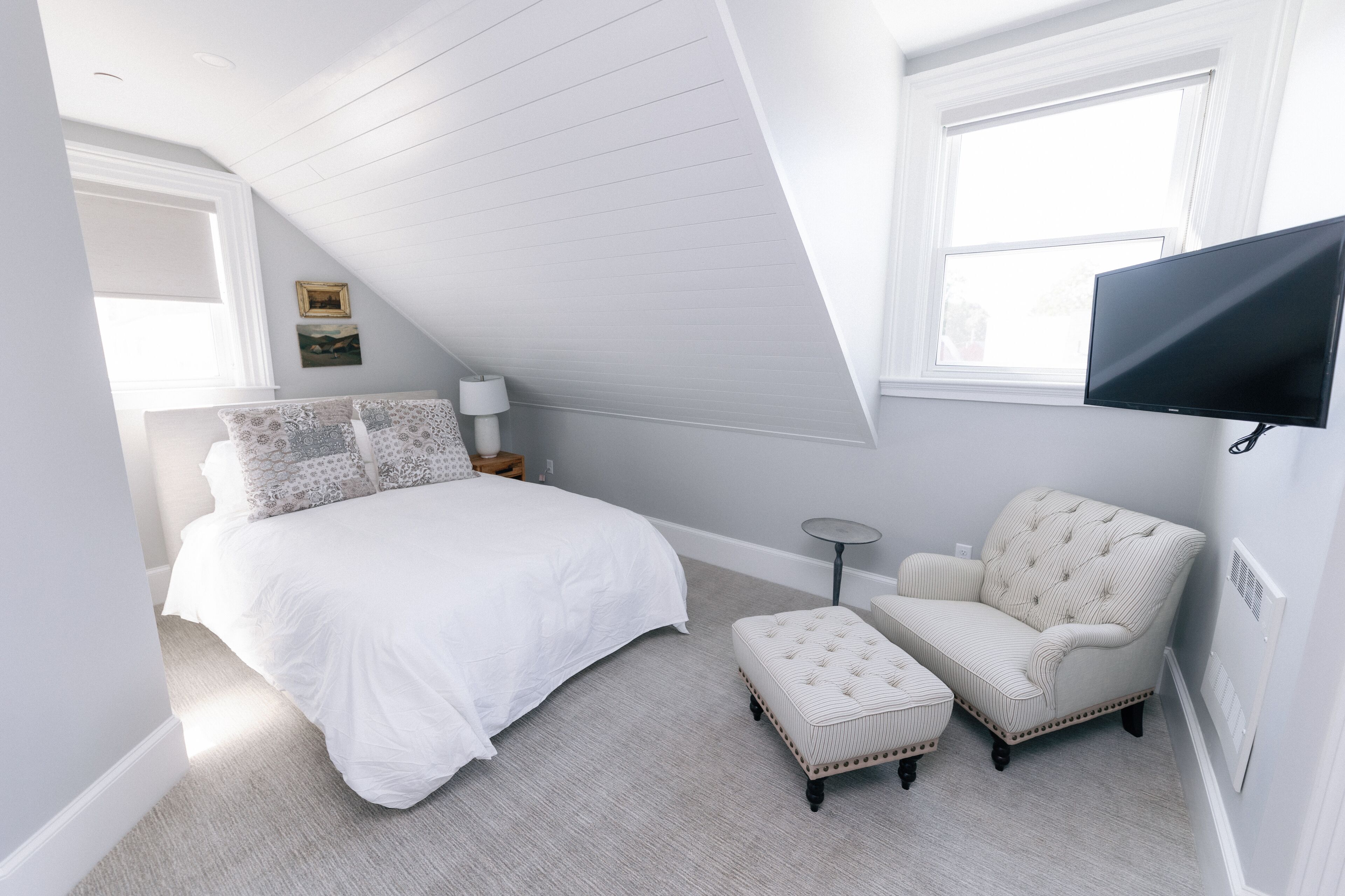 A bedroom with a sloped ceiling, a bed with white bedding, a chair, a small table, and a TV mounted on the wall.