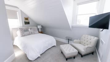 A bedroom with a sloped ceiling, a bed with white bedding, a chair, a small table, and a TV mounted on the wall.