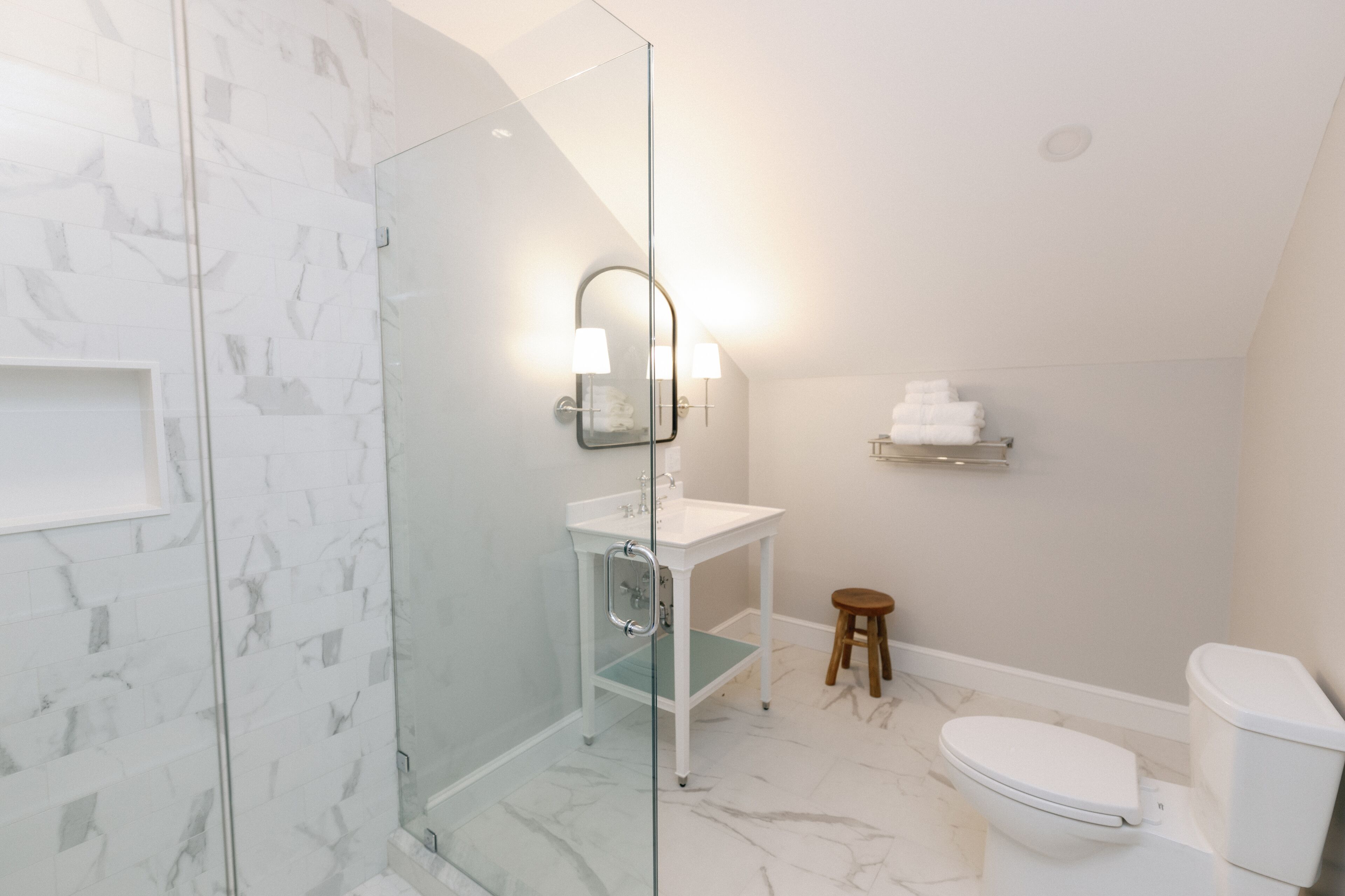 A modern bathroom with a glass shower enclosure, a white sink, a mirror with lights, a toilet, and a small stool.
