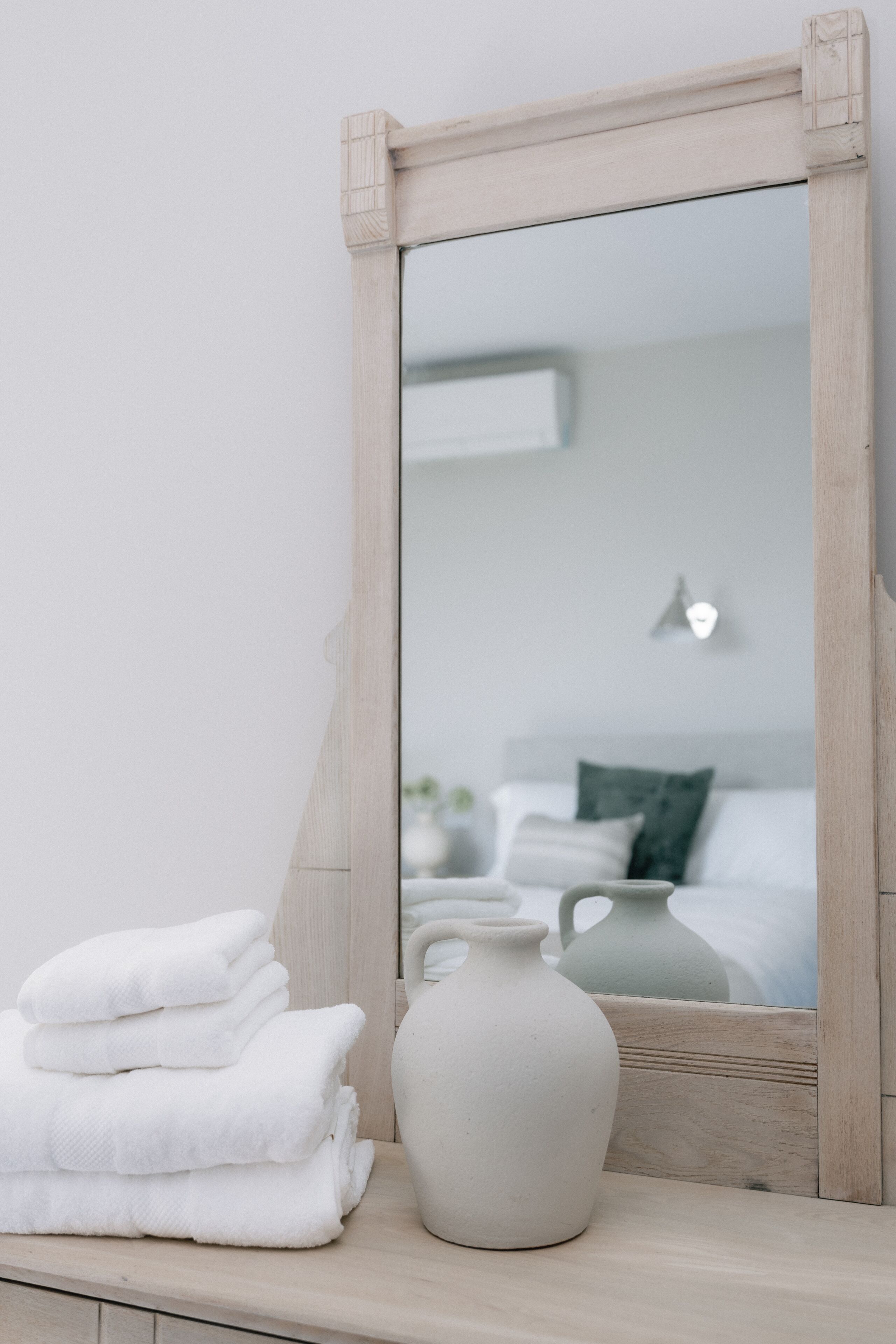 Een spiegel met houten lijst, een stapel witte handdoeken en een witte vaas op een houten oppervlak.