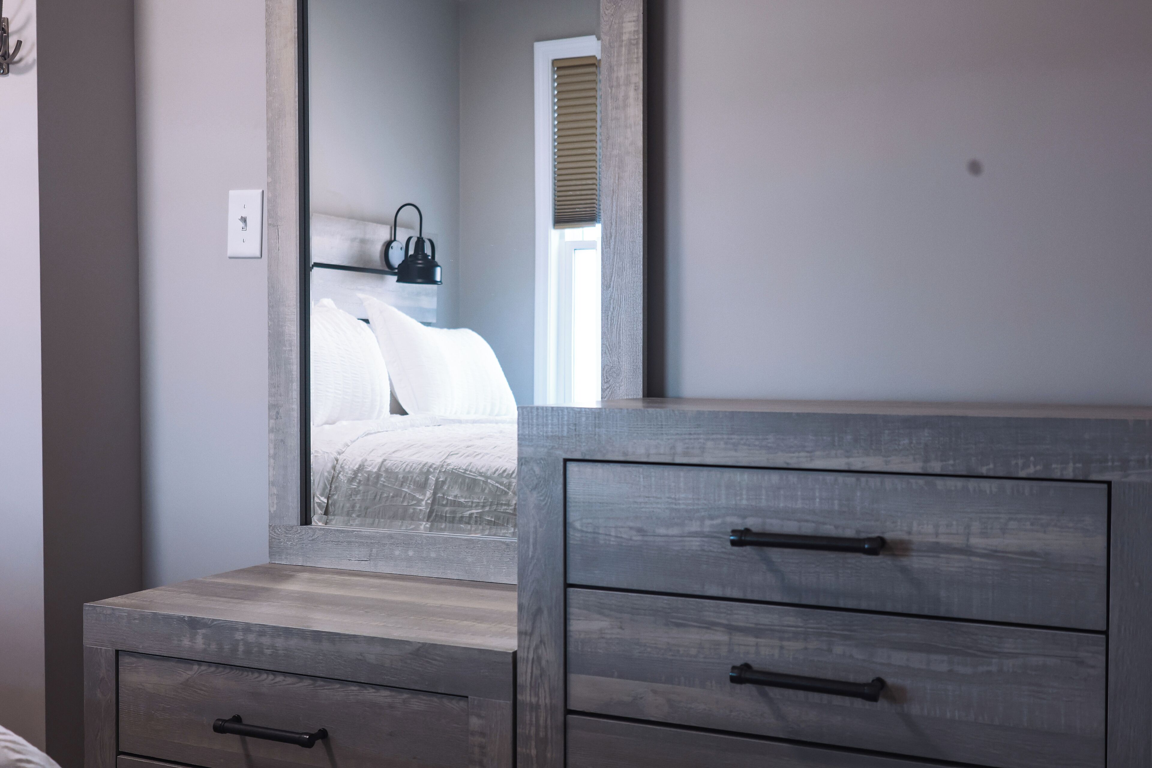 A bedroom with a large mirror, wooden dresser with drawers, a bed with white bedding, and a window with blinds.