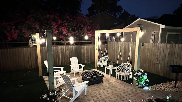 Ein Hinterhof mit einer Terrasse, weißen Stühlen, einer Schaukel und einem kleinen Tisch.