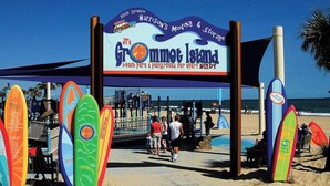 Ein Zugang zum Strand mit einem Schild für Grommet Island, zeigt Surfbretter und einen Strandabschnitt.