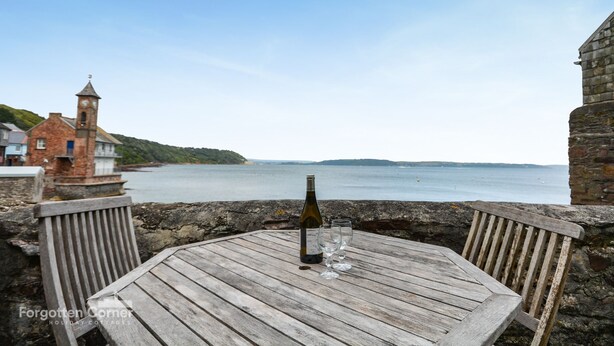 Una mesa de madera con una botella y vasos, con vista al mar tranquilo y un edificio histórico con una torre del reloj.