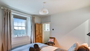 Un dormitorio con una cama, una ventana con cortinas, una puerta de madera, una pequeña mesa con una bandera y una lámpara en el techo.