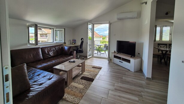 Una sala de estar moderna con un sofá de cuero, una mesa de centro y un televisor sobre un mueble. Hay un ventanal corredizo que da a un balcón con mobiliario exterior.