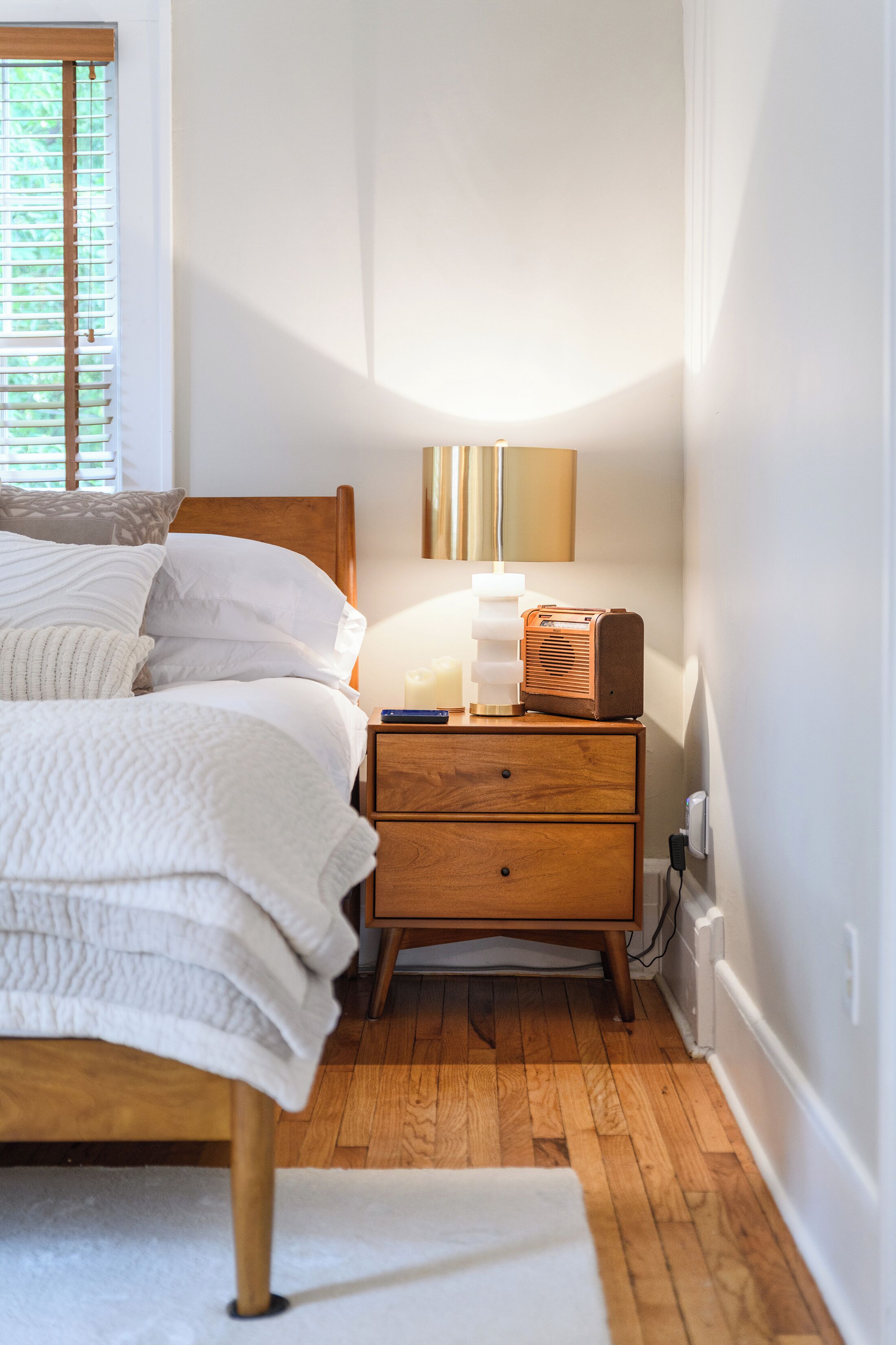 A neatly made bed with white bedding, a wooden nightstand with drawers, a lamp, and a vintage radio.