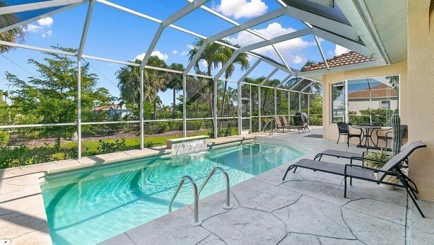 A swimming pool with a glass enclosure, lounge chairs, and a patio area with a table and chairs. - Mile From Beach Access, Spacious and Redone, Heated Pool! (Marco Island)