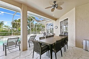 A spacious indoor dining area with a large table and chairs, a ceiling fan, and large windows offering a view of the outdoors.